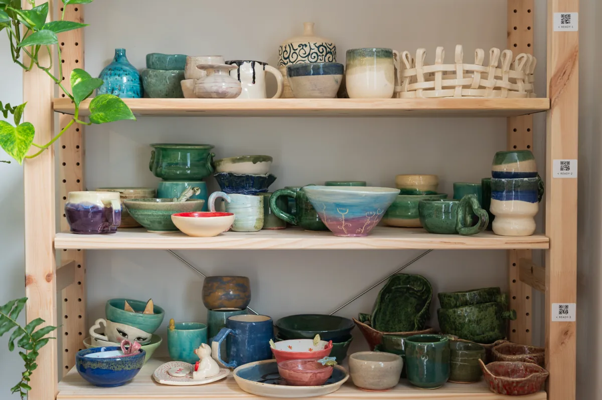 Shelves of glazed student pieces: mugs, bowls and vases in greens, blues and pinks